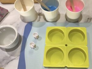 three paper cups filled with white, blue and pink melted soap base sitting next to a breadboard with a four-cavity circular silicone mould sitting on it