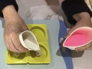 pouring white melted soap base from a paper cup into a paint pouring tool sitting in the centre of a circular silicone mould cavity