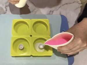 pouring pink melted soap base from a paper cup into a paint pouring tool sitting in the centre of a circular silicone mould cavity
