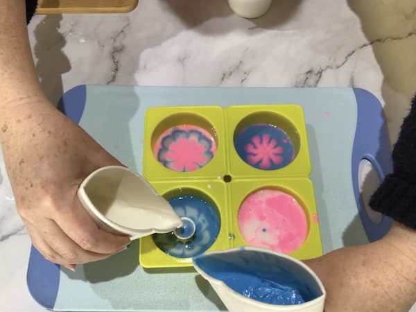 pouring white and blue melted soap base from a paper cup into a paint pouring tool sitting in the centre of a circular silicone mould cavity