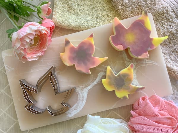 three autumn leaf melt and pour soap bars sitting on a pink marble resin board with three nested maple leaf cookie cutters