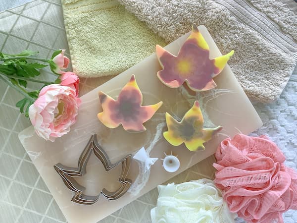 maple leaf cookie cutters, acrylic paint pouring tools and autumn leaf soaps laid out on a pink resin board surrounded by face washers and loofas