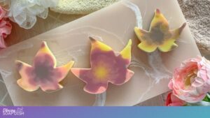Three leaf shaped soaps with soft blended colours of pink, yellow, and purple in a tie dye pattern rest on a marble surface. Flowers and a folded towel surround them, creating a gentle spa inspired arrangement.