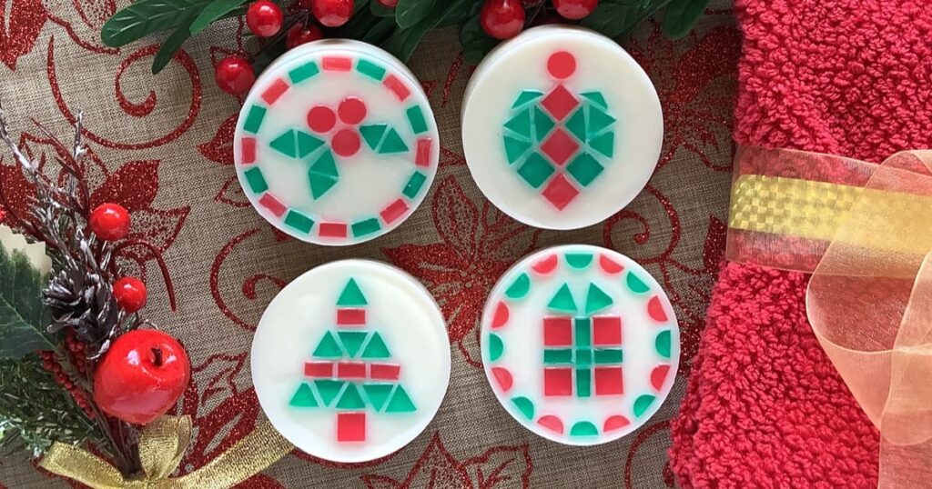 Four round, white soaps decorated with red and green mosaic tile Christmas designs sit on a festive red and gold cloth. The patterns include holly leaves and berries, a Christmas tree ornament, a stylized Christmas tree made of triangles, and a wrapped gift with a bow. Holiday greenery, red berries, and a gold tied red towel surround the display.