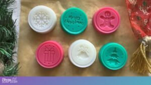 Six round soaps with Christmas designs are arranged on a brown surface. The top row shows a white soap embossed with a snowflake design, a green soap embossed with the text “Merry Christmas”, and a red soap embossed with gingerbread man figure. The bottom row features a red soap with an embossed image of a gift, a white soap with an embossed image of a bell, and a green soap with an embossed image of a Christmas tree. The soaps have specs of glitter in them, and they are surrounded by festive greenery and red fabric accents.
