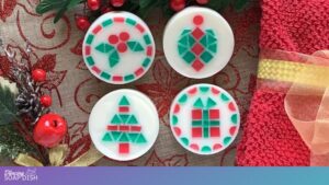 Four round white soaps with embedded red and green mosaic tile Christmas designs are arranged on a festive surface. The patterns include holly leaves, an ornament shape, a Christmas tree, and a gift box. Red ribbon, gold accents, berries, and greenery surround the display, creating a bright holiday setting.