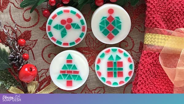 Four round white soaps with embedded red and green mosaic tile Christmas designs are arranged on a festive surface. The patterns include holly leaves, an ornament shape, a Christmas tree, and a gift box. Red ribbon, gold accents, berries, and greenery surround the display, creating a bright holiday setting.
