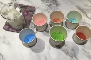 six paper cups with cookie stamps inside sitting next to a jug of melt and pour soap base