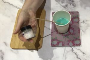 green melt and pour soap base in a paper cup with a had holding a jar of glitter