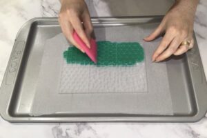 a hand holding a scraper scraping green soap base across the surface of a mosaic tile silicone mould