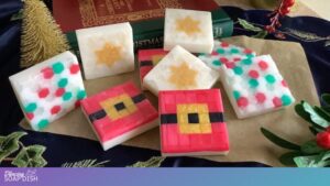 Square handmade soaps with festive mosaic tile designs sit on brown paper accented by a dark blue cloth and holiday décor. The soaps feature patterns like Santa belt stripes, gold stars, and colourful red, green, and white glass tile mosaic. Behind them are red and green books that add to the Christmas theme.