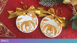 Two round Christmas ornaments rest on a red glittery surface, each featuring a gold paint stamped nativity scene with Mary, Joseph, and baby Jesus beneath a star. Gold hanging ribbons, a wire tree decoration, and green pine branches surround the festive display.