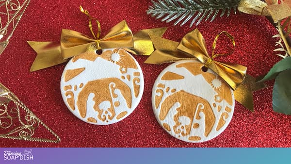 Two round Christmas ornaments rest on a red glittery surface, each featuring a gold paint stamped nativity scene with Mary, Joseph, and baby Jesus beneath a star. Gold hanging ribbons, a wire tree decoration, and green pine branches surround the festive display.