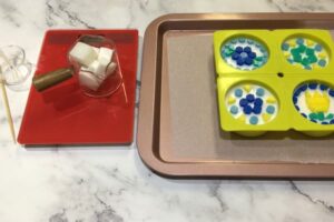a small glass measuring cup filled with white soap base cubes sitting next to soap bars with mosaic soap tile designs on top