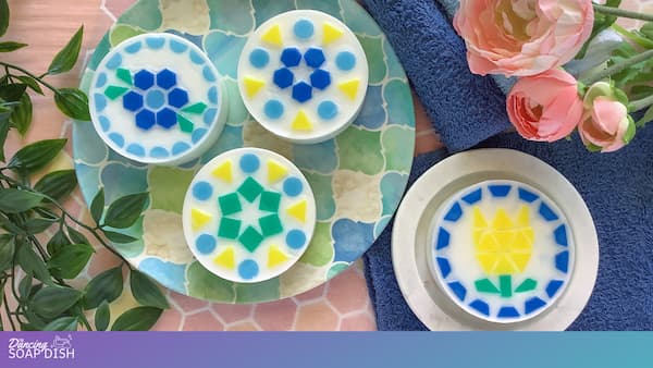 Four round soaps with mosaic style patterns in blue, yellow, and green sit on a patterned plate and a white dish. Each soap features a different geometric floral or leaf design. A blue towel, pink flowers, and green leaves surround the arrangement, creating a colourful, spa like display.