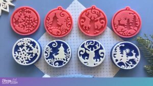 Four pink silicone moulds with winter designs sit in the top row, including snowflakes, snow scenes, and reindeer motifs. Below them are four round blue and white soap bars showing the same patterns in reverse relief. A light blue background, textured white fabric, and a small pine sprig complete the festive craft setup.