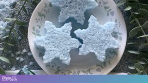 Three light blue star shaped Epsom salt cakes with a crystalline, snowflake like texture sit on a white plate decorated with green leaves. Bath salts and small green sprigs are scattered around the plate, creating a fresh, spa style scene.