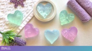 Translucent heart shaped glycerine soaps in soft pastel shades of green, blue, and pink are arranged on a light surface. One sits in a white dish while others rest nearby. Different cracked ice effects are present in all the bars, making them look as if they are frozen. A textured white towel, two rolled lavender towels, and sprigs of lavender flowers create a calm, spa like setting around them.