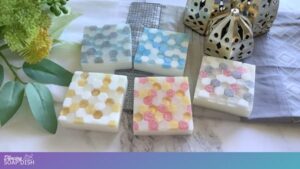 Five square handmade soaps with colourful hexagonal mosaic tile patterns sit on a marble surface. The patterns on the surface of the soaps emulate decorative glass mosaic bathroom tiles. Green leaves, a yellow flower, a small metallic lantern, and folded flannel cloths surround them, creating a decorative, artisanal display.