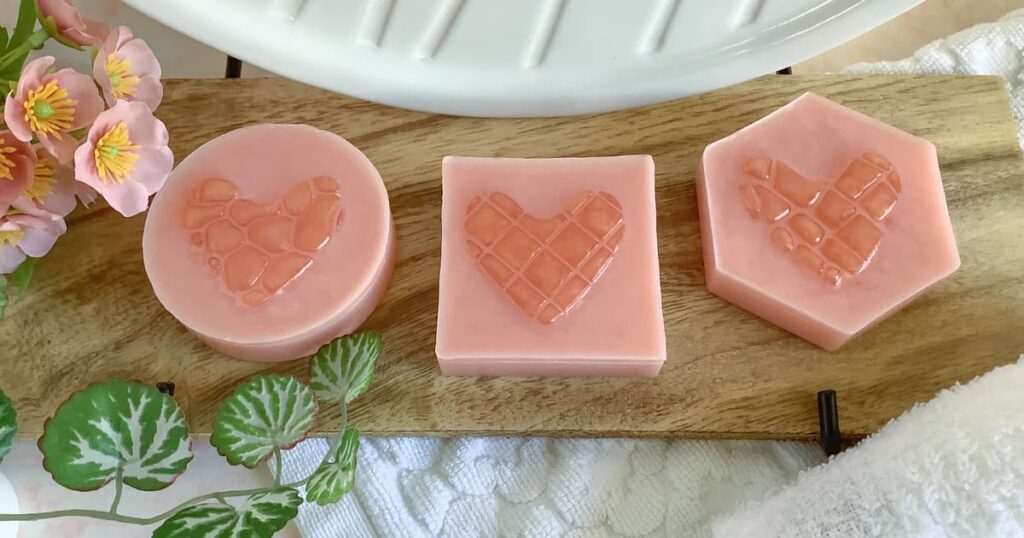 Three pink handmade soaps—a round, square, and hexagonal bar—sit on a wooden tray, each featuring a raised heart design with a textured mosaic pattern. Pink flowers, green leaves, a white towel, and a white soap dish surround the display, creating a soft, decorative scene.