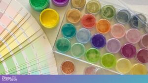 Small round containers filled with brightly coloured mica pigment powders—greens, yellows, reds, oranges, blues, purples, and pinks—sit neatly in a clear tray. Beside them, a fan of pastel colour swatch cards spreads outward, creating a vibrant, craft focused display.