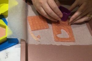 a pair of hands using a cookie cutter to cut a heart shape from a slab of mosaic soap tiles