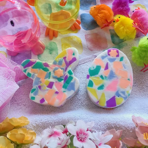 A pastel Easter‑themed display featuring a bunny‑shaped soap and an egg‑shaped soap decorated with colorful mosaic pieces in pink, yellow, green, purple, and blue. Around them sit fuzzy chick decorations in bright yellow, orange, and purple, along with translucent animal‑shaped containers. The background includes a soft white towel with watercolor spots and pink and yellow artificial flowers, creating a cheerful springtime scene.