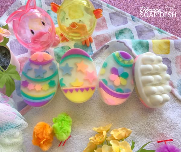 Four Easter egg shaped massage bar soaps decorated with various geometric patterns are arranged on a colourful tea towel. The last bar on the right is overturned to show the rounded nodules on the bottom of the massage bar. Small Easter decorations surround the display.