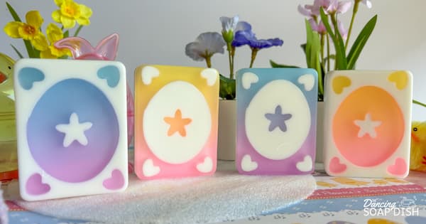 Four rectangular pastel soaps with Easter egg designs in the centre and small hearts in each corner are arranged on a soft towel. The soaps feature purple to blue, and yellow to pink gradients. Colourful spring flowers in yellow, purple, and pink sit in the background, and the corner includes a ‘The Dancing Soap Dish’ watermark.