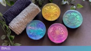 Four round translucent soaps in bright colours—yellow, green, blue, and pink—are arranged on a dark surface surrounded by green leaves. Each soap is embossed with an intricate floral or mandala style pattern. Two folded grey towels sit beside them, creating a calm, spa like display.