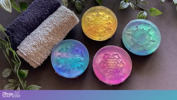 Four round translucent soaps in bright colours—yellow, green, blue, and pink—are arranged on a dark surface surrounded by green leaves. Each soap is embossed with an intricate floral or mandala style pattern. Two folded grey towels sit beside them, creating a calm, spa like display.