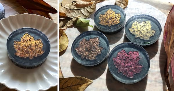 A round soap with a metallic gold maple leaf sits on a white scalloped dish, while four additional soaps—each featuring a maple leaf in gold, bronze, copper, and red—are arranged together on a dark surface. Each leaf has small, round dew drops on them. Dried autumn leaves surround the soaps, creating a warm, seasonal display.