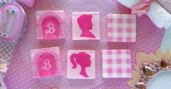 Six ‘Barbie Movie’ themed square soap bars are arranged on a pink polka dot background, each featuring a different design: Barbie Dreamhouse, a Barbie profile silhouette, and a pink and white checkered fabric pattern, repeated in two rows. Decorative items such as a jeweled case, fluffy white fabric, and a crown like accessory with pink gemstones surround the display.