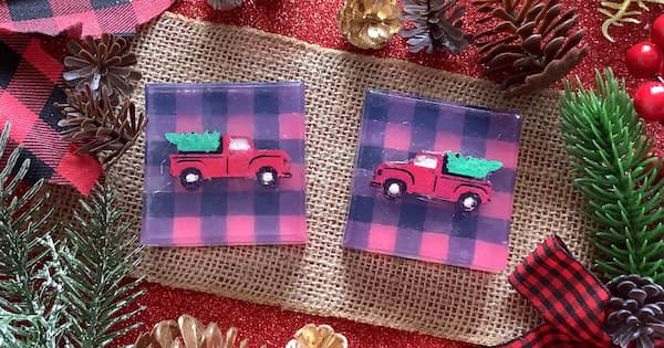 Two square soap bars with embedded red vintage pickup trucks carrying green Christmas trees sit on burlap fabric. Each soap has a red and black buffalo plaid background. Pinecones, evergreen branches, red berries, gold ornaments, and a matching plaid ribbon surround the scene, creating a rustic holiday display.