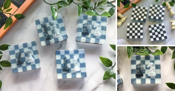Several square black‑and‑white chess-themed soaps sit on a marble surface. The front of the soap diplays two chess pieces under a translucent layer. The back of the soap displays a black and white checkerboard pattern. Green leaves surround the display, and a few loose miniature chess pieces appear nearby.