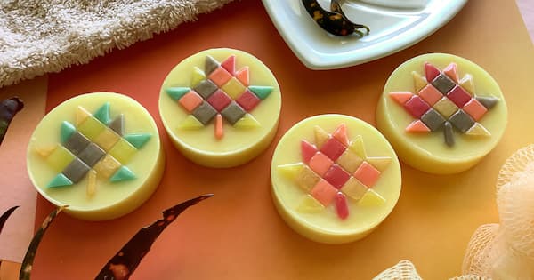 Four round handmade soaps are arranged in a row, each topped with an intricate geometric pattern in the shape of a maple leaf made of small, square soap tiles in shades of pink, yellow, green, gray, and orange. A white towel, a dish with a decorative spoon, and other bath accessories sit in the background, creating a calm, spa‑like setting.