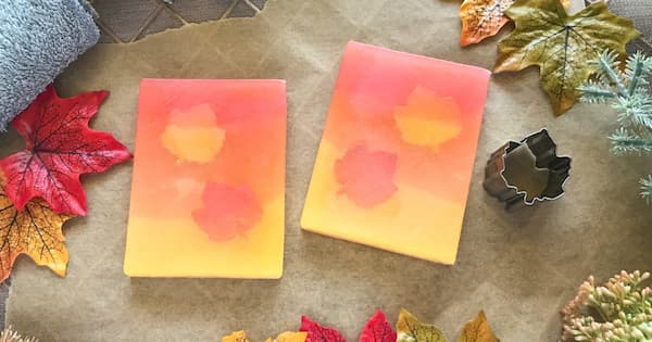 Two rectangular handmade soaps with a pink‑to‑orange‑to‑yellow gradient sit on parchment paper, each featuring a subtle leaf design in the center. They are surrounded by colorful autumn leaves, a metal leaf‑shaped cutter, textured fabric, and greenery, creating a warm fall‑themed arrangement.