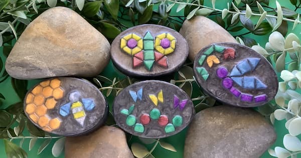Five black, oval soap bars decorated with bright geometric mosaic designs resembling insects rest among larger natural rocks and green leaves. The patterns include a butterfly, a bee, a dragonfly, and a caterpillar design made from colorful mosaic soap tiles in the shape of triangles, hexagons, circles, and squares.