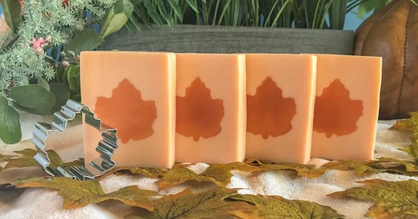Four rectangular handmade soap bars in a soft orange color are arranged in a row, each featuring a darker orange maple leaf embedded in the center. A metal maple‑leaf cookie cutter sits in front of them, surrounded by artificial greenery and scattered autumn leaves on a light fabric background, creating a cozy fall-themed scene.