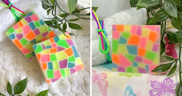 Two square handmade soaps with bright neon mosaic pieces in pink, orange, green, yellow, purple, and blue sit on a textured white surface with green leaves around them. On the right, a single upright soap with the same colorful geometric pattern is displayed near a white towel and greenery.