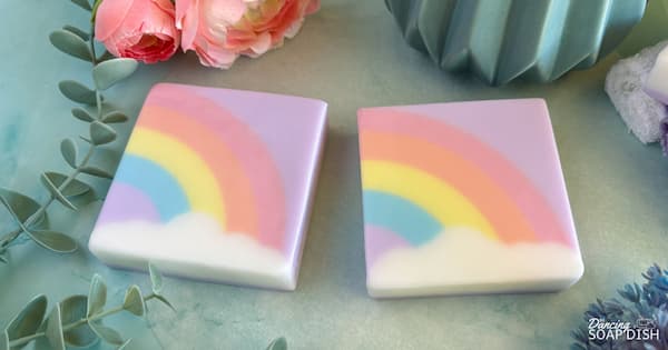 Two square pastel soaps with arched rainbow designs of pink, orange, yellow, green, and blue above soft white clouds with a lavender-coloured background, rest on a light bluey-green surface. Pink flowers, green leaves, and a teal ceramic container surround them, creating a bright, cheerful display.