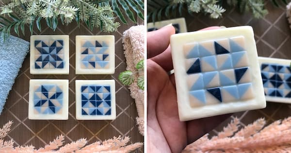 Four square blue, white and cream geometric soaps are arranged on a textured brown surface with soft fabrics and greenery around them. A close‑up on the right shows one soap being held in a hand, highlighting its detailed quilt block mosaic pattern.