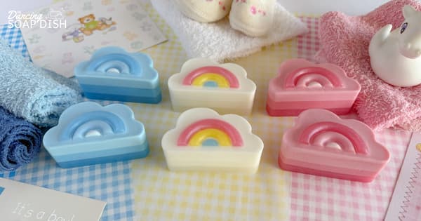 Six cloud‑shaped soaps with tiny rainbow embeds in pastel blue, pink, and white are arranged on a soft pastel gingham surface. Around them are folded blue and pink towels, baby socks, and greeting cards, creating a gentle, gift‑like display.