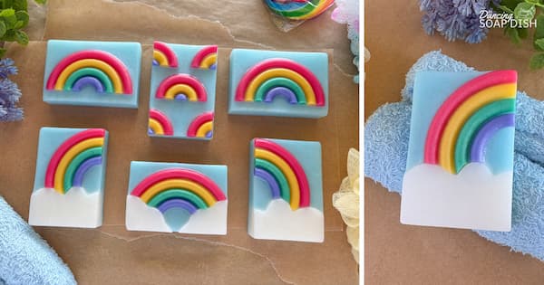 Several rectangular soaps with various bright rainbow designs are arranged neatly on a brown-paper surface with towels and flowers nearby. Each soap bar has a pale blue background, and the designs include single arched rainbows, rainbows rising from soft, white cloud shapes, and a mini rainbow repeating pattern. A close up on the right shows one bar resting on a blue towel.
