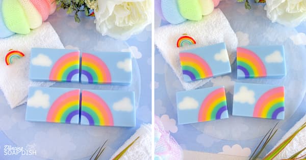 Four rectangular soaps with a rainbow‑and‑cloud design are arranged on a light blue cloud‑patterned surface. Each bar shows a white cloud on one side with a rainbow arcing across a pale blue background. On the left, the bars are grouped to form two complete rainbows; on the right, the same bars are separated to show each individual piece. White towels, pastel bath sponges, flowers, and a small rainbow decoration surround the display.