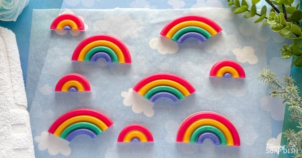 Several rainbow shaped soap embeds with bright coloured layers of red, yellow, green, and blue are arranged on a light blue surface patterned with soft white clouds. The rainbows are various shapes and sizes, some full arches and some partial arches.  There are also some very tiny three-layer rainbow soap embeds. A towel sits to the left and green plants to the right, creating a fresh, cheerful display.