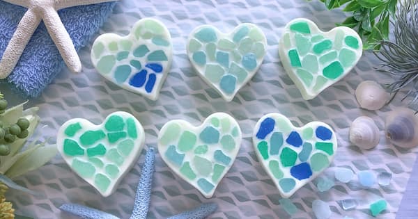Six white, heart shaped soap bars are arranged in two rows on a lightly patterned surface. Each heart features a mosaic pattern of sea glass like geometric fragments on its surface in shades of blue, turquoise, and green. Starfish, seashells, and green plants surround the display, creating a coastal, beach themed scene.