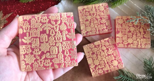 A hand holds a square red soap bar embossed with detailed Christmas motifs: snowflakes, reindeer, ornaments, candy canes, and gift boxes. Three matching soaps rest on a beige surface in the background, surrounded by pine branches and festive red décor.