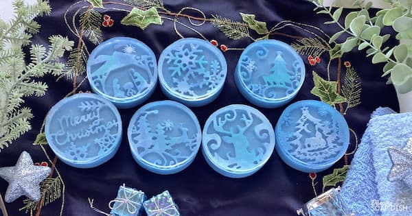 Seven round blue soaps with detailed winter and Christmas designs including a nativity scene, a snowflake, a Christmas tree, “Merry Christmas,” a reindeer, and a snowy village. They are arranged on dark fabric decorated with a holly motif, and surrounded by green foliage and silver and blue ornaments.