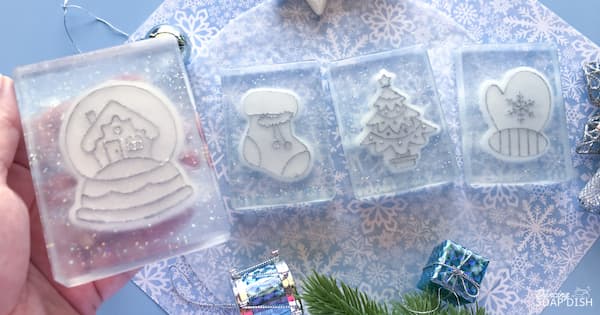 Four transparent rectangular soaps with white winter designs embossed with sparkly silvery deatails, a snow globe with a house, a Christmas stocking, a decorated Christmas tree, and a mitten with a snowflake, are arranged on a light blue snowflake‑patterned surface. Small wrapped gifts and pine branches surround the display, creating a festive holiday scene.